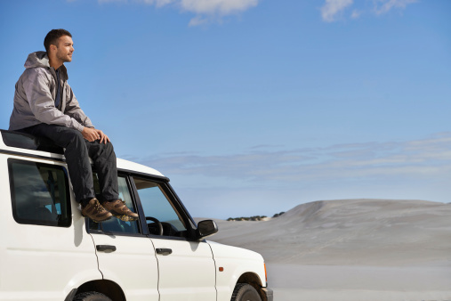 Cropped shot of a young man sitting on the roof of his car while on a roadtrip