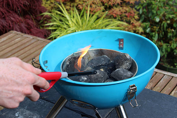 Lighting charcoal barbecue / BBQ lighter / flames on kettle barbecue coals Photo showing a person lighting the charcoal briquettes / coals with a long BBQ lighter tool.  This portable kettle camping barbecue is pictured on a wooden garden table, on protective slate placemats, and has been lit with the aid of small barbecue fire lighters. Aflame stock pictures, royalty-free photos & images