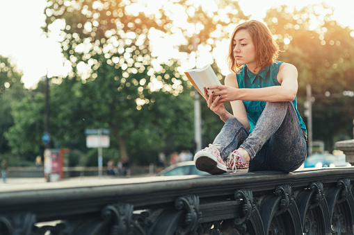 Teenage girl reading a book while sitting on a bridge near big boulevard.