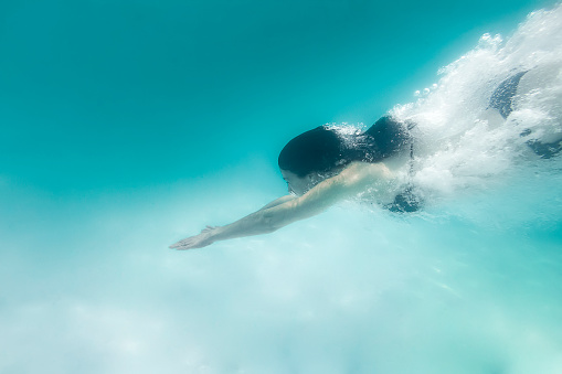 A DSLR underwater photo of a 35-year-old  caucasian beautiful girl diving in clear blue water in Rio de Janeiro, Brazil. She has just jumped into the water with her stretched armys in front of her. She is wearing a black bikini. She is covered with bubbles created by the dive.