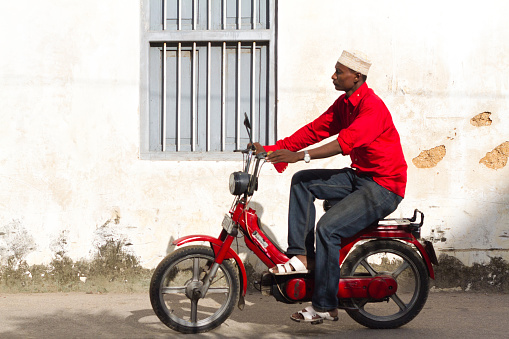Stone Town (Zanzibar), Tanzania - May 22, 2012: Man in a red shirt and white skull cap riding a red scooter near a white wall in Stone Town, Zanzibar.