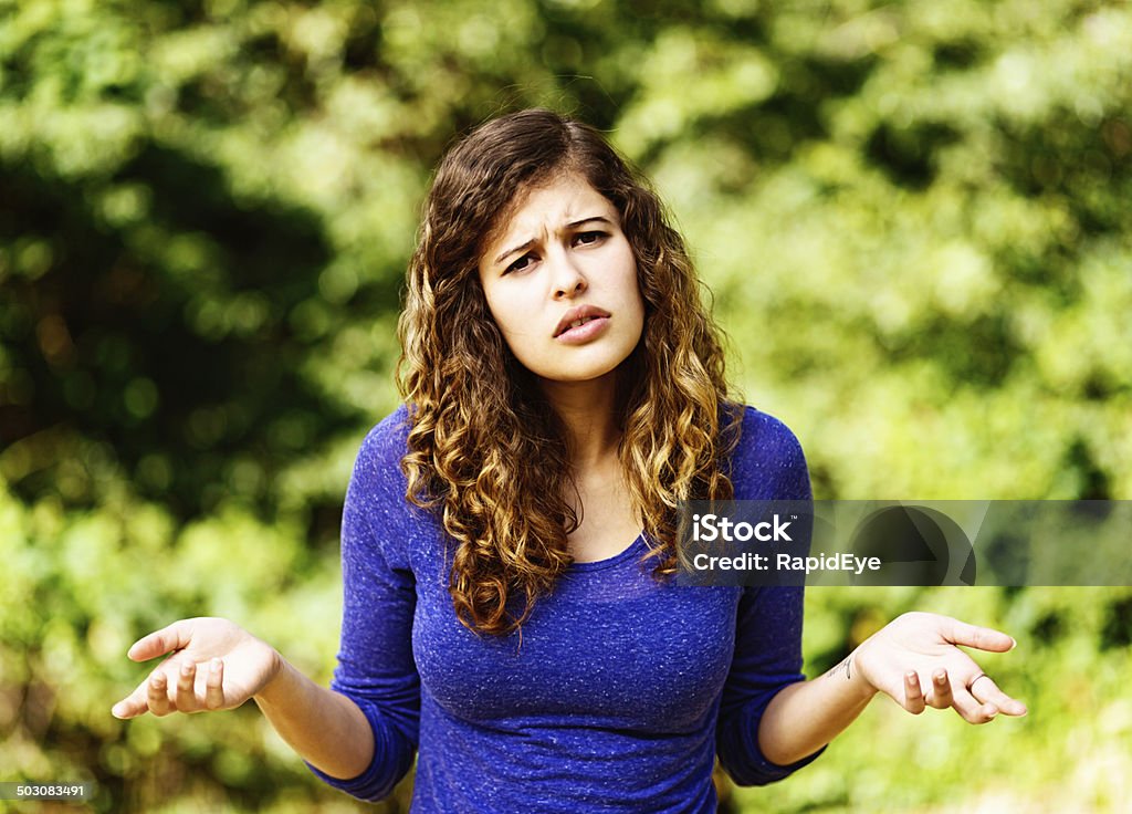 Frowning young woman shrugs in irritation against foliage This beautiful young honey-blonde, brown-eyed woman frowns and shrugs against a background of greenery, irritated and confused. One Woman Only Stock Photo