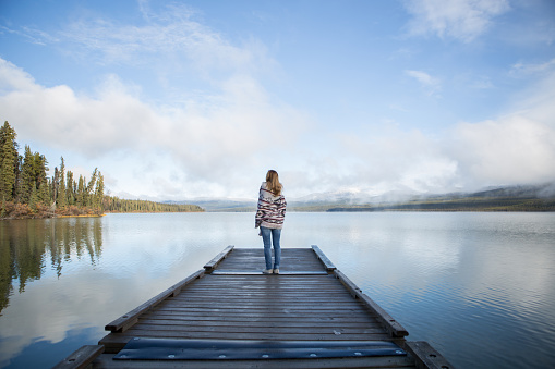Young woman stands on wooden pier in Canada looking away. She is wearing a poncho.  Morning sunlight passing through the mist.