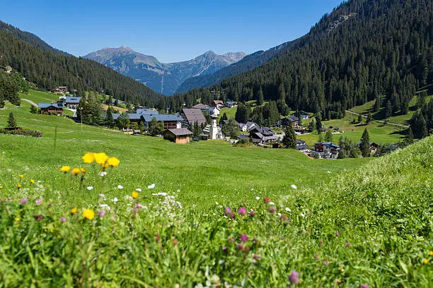 Pretty village in the Alps, Austria, on a summer day. Green grass with flowers in the foreground, a church, wooden houses and village in the background. In the distance you can see mountains, the Alps with fir trees.