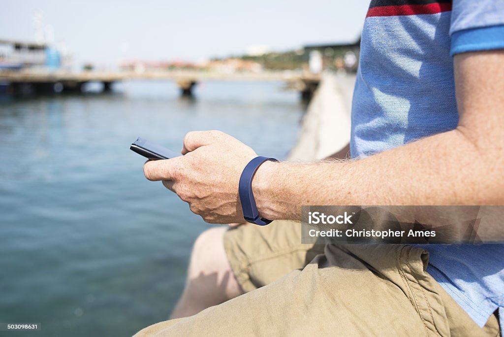 Getting Healthy with Activity Wristband A man sitting by the sea, wearing casual sports clothes using a smartphone and activity tracking wristband. Activity trackers can communicate with smart phone applications and are designed to help the wearer achieve a more active and healthy lifestyle. Activity Stock Photo