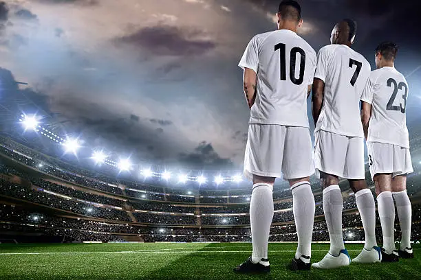 Defensive wall of football players standing on pitch during game in full floodlit stadium under stormy sky