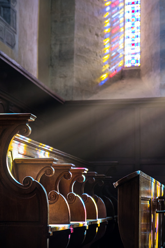 Vertical composition photography of sunbeam through colored stained glass in french church of medieval city of Cremieu in Isere, Rhone-Alpes region in France (Europe). There are several wooden sculpted pews seat in choir space in a row illuminated by sunlight. Those church seat (stalls), especially one next to the chancel or choir, are reserved for church officials and dignitaries.
