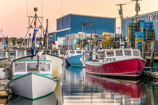 Photo of colourful fishing boats in harbour at sunset. The boats are tied up to wooden jetties. Piles of colourful metal lobster traps along with other nautical equipments are visible in background. Portland, Maine.