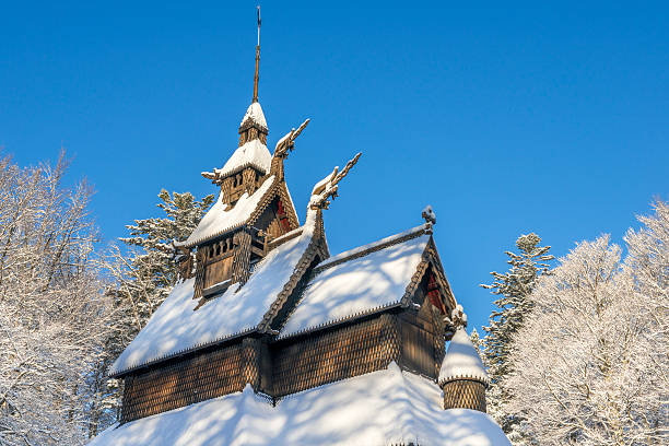 Winter landscape with medieval wooden Fantoft Stave Church in Bergen on the West Coast of Norway with snow-covered roof after the first snowfall of the season. In the background are the tall trees. The branches are covered with snow. The sky is clear and blue.