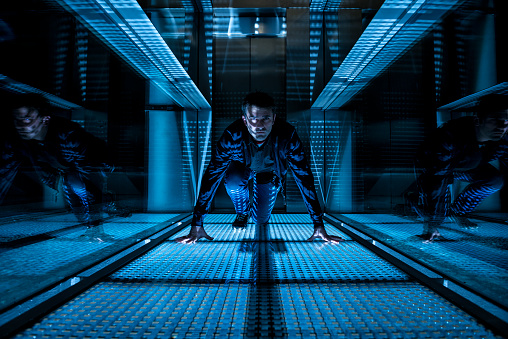 At night a mature man (maybe a special agent or a technician) is crawling on the floor of a corridor with an elevator in the background. He is wearing a blue jean and a shirt with long sleeves and he is looking concentrated at the camera. He is in a starting position with his hands down at the floor.