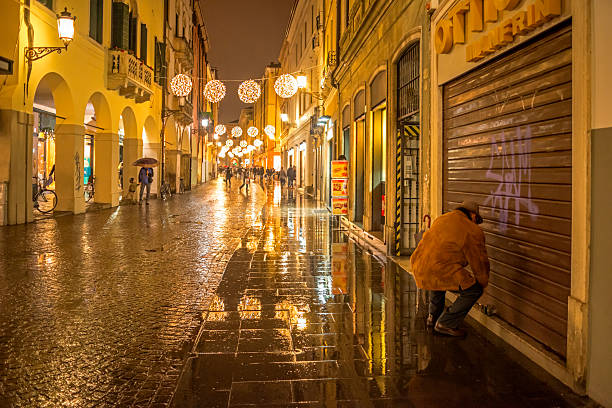 Padua, Italy - January 4, 2014: Shop owner squating down for locking the window display of his shop on the rainy January evening, Rain just washed the streets of old Padua, Italy. Visible and recognisable faces of the pedestrians. Shop names, advertisements and brands visible down the street. Pedestrians walk by in the distance.