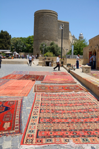 Baku, Azerbaijan - July 4th, 2014: Maiden Tower viewed from Asef Zeynalli street in a sunny day. Many carpets for sale lying on the street in the foreground. Some people walkin on the square. Vertical image.