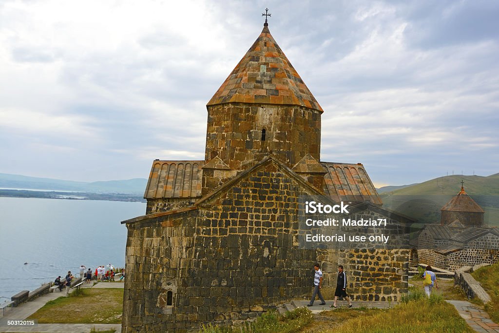 Sevan Sevanavank, Armenia - July 6th, 2014: Sevanavank monastery complex viewed from the top of the hill. In the middle the main church, the chapel on the right. On the left some people on the terrace and Sevan lake. Two men and a woman walking around the church. Horizontal image in a cloudy day. Architecture Stock Photo