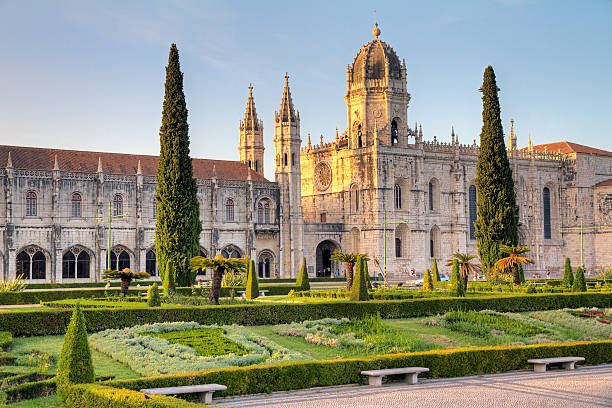 Ornate stone monastery with towers and spires, fronted by formal gardens with cypress trees in golden sunlight.