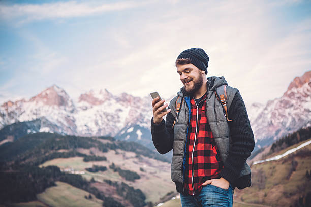 Healthy lifestyle A man on vacation hiking in Austrian Alps. Wearing warm clothes and backpack. He took a break to rest, check his smart phone and enjoy in beautiful landscape. Winter day. Mountains are covered with snow. extreme terrain stock pictures, royalty-free photos & images