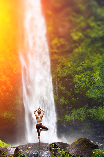 A young woman practicing yoga outdoors in front of the beautiful waterfall.