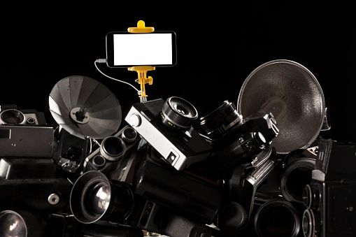 Modern smartphone placed on a selfie stick on top of old fashioned cameras.Large amount of old fashioned cameras are placed on bottom of frame.The light is placed on back.Shot with a full frame DSLR camera in studio.The screen of smartphone is blank white.