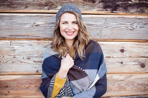 Woman on vacation at mountain cabin. Standing in front of house by wooden wall and enjoying in winter day. Wearing warm clothing, hat and scarf. Austrian Alps.