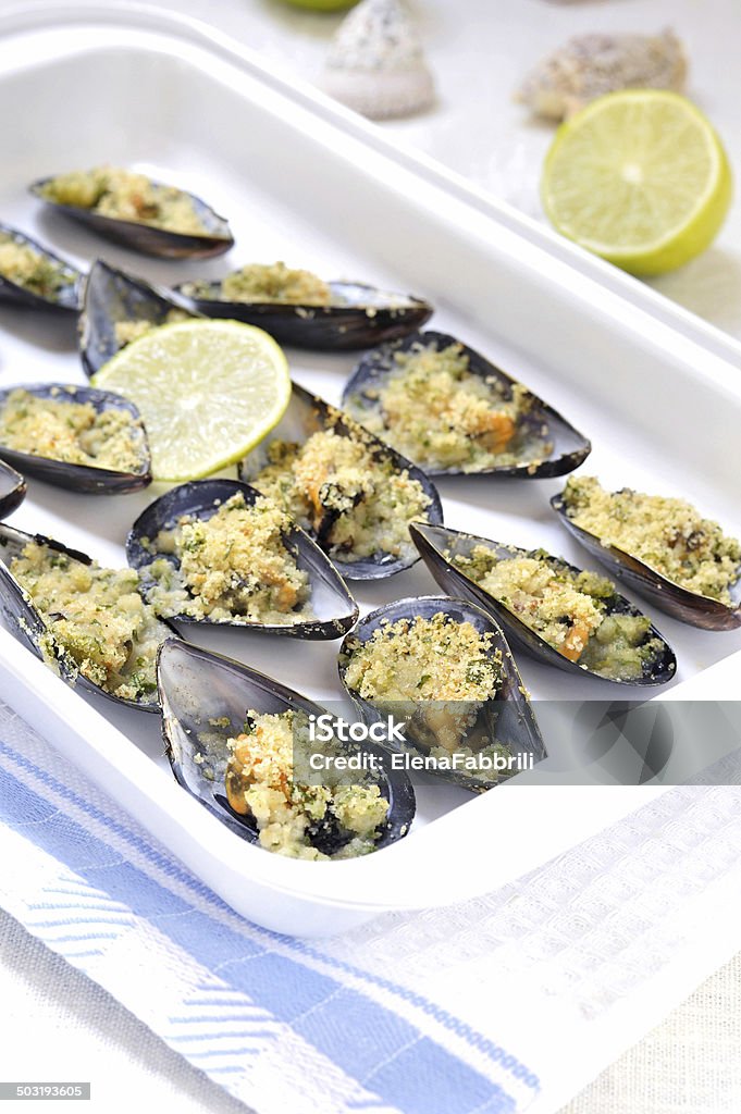 Gratin of Mussels, selective focus. Italian cuisine. Italian appetizer. Baked fresh mussels under the bread crumbs on a white enamel baking tray, decoration - seashells, lemon. Animal Shell Stock Photo