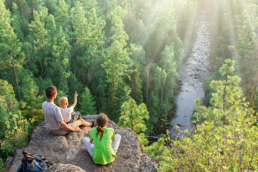 Hikers with backpack sit on big rock above green forest and river
