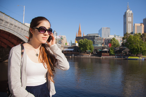 A young woman on her mobile phone. She is sitting next to the Yarra River in Melbourne, Australia with the central city area visible behind her. Copy space.