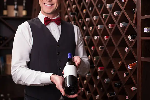 I have a special drink for you. Skillful young sommelier is holding a bottle of red wine. He is standing in cellar and smiling