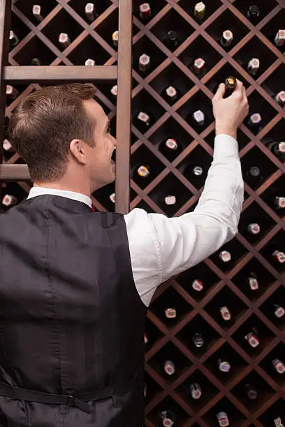 Professional young sommelier is choosing a bottle of wine in a cellar. He is standing in a tuxedo. The man is smiling happily