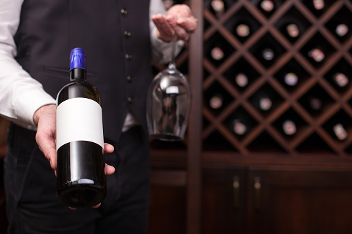 Close up of hands of young sommelier presenting a bottle of red wine. He is standing in cellar and holding a glass. Copy space in right side