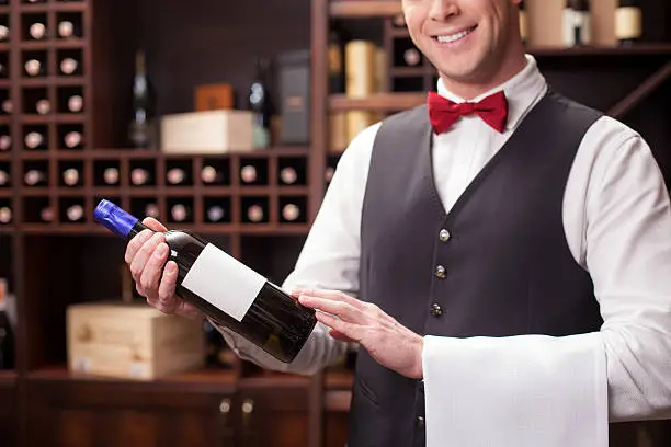 Professional male wine waiter is serving a customer. He is standing and holding a bottle of red wine. The man is carrying a towel and smiling