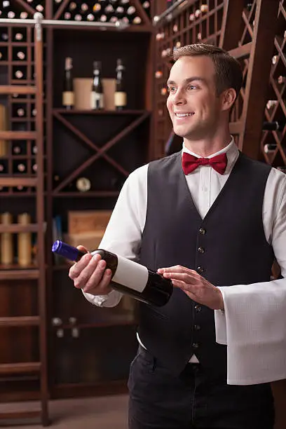 Handsome young sommelier is carrying a bottle of red wine in cellar. He is standing and smiling. The man is looking aside with anticipation