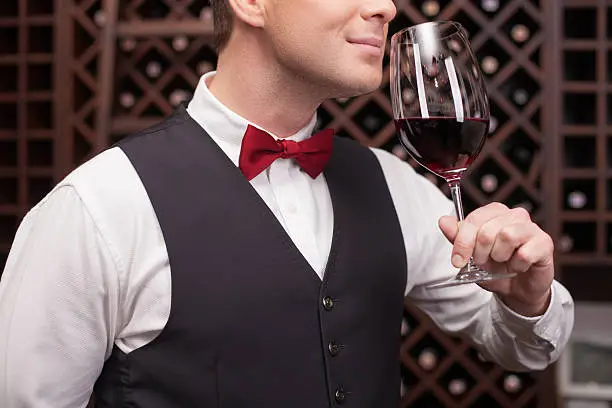 Professional young sommelier is smelling wine with enjoyment. He is holding a wineglass near his nose. The man is standing and smiling