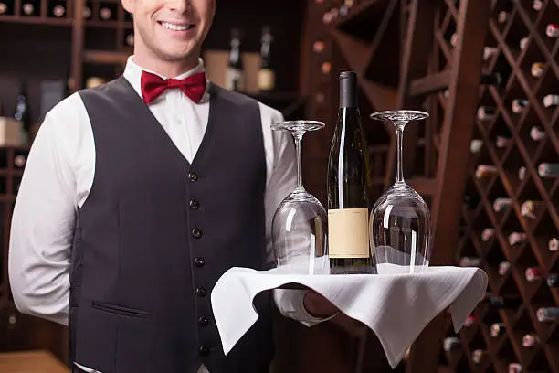 Skillful young sommelier is serving a bottle of wine and wineglasses to the customer. He is standing and holding a tray. The man is posing and smiling