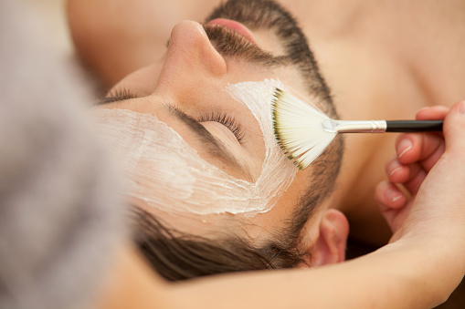 Close up of attractive man lying and relaxing at spa. The beautician is applying facial cream on his skin