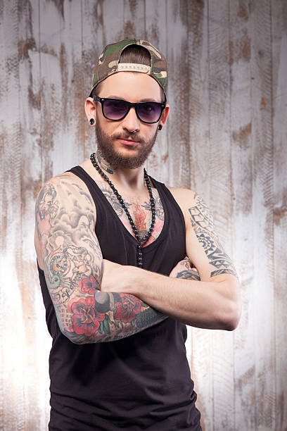 Attractive bearded guy is expressing his confidence Handsome young hipster with tattoo and beard. He is standing and crossing his arms. The man is standing and looking at camera confidently cross shoulder tattoos stock pictures, royalty-free photos & images
