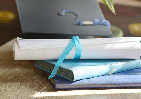 A mortarboard and graduation scroll, tied with blue ribbon, on a stack of old battered book with shallow depth of field