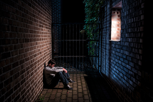 A mature man with beard (maybe a homeless, a refugee,...) sitting alone outside on the ground. It is at night and he is sitting in a light beam from the illuminated window on the opposite side. His posture shows sadness, depression and loneliness.