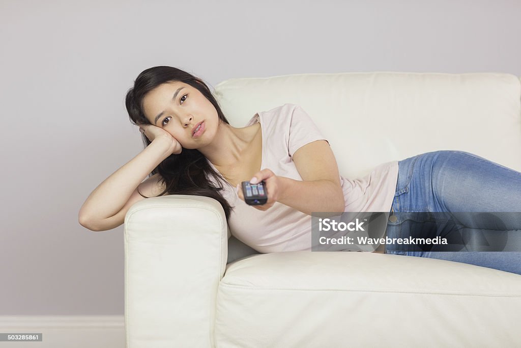 Bored asian girl lying on the couch watching tv Bored asian girl lying on the couch watching tv at home in the sitting room Holding Stock Photo