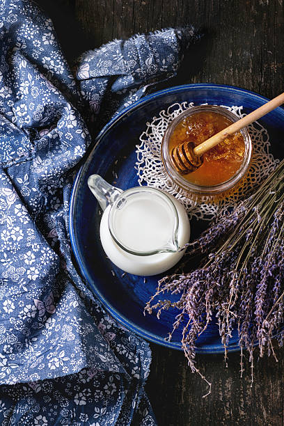Honey, milk and lavender Open glass jar of liquid honey with honeycomb and honey dipper inside, jug of milk and bunch of dry lavender in blue plate over old wooden surface with blue textile rag. Dark rustic style. Top view blue jug stock pictures, royalty-free photos & images