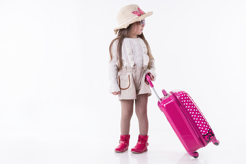 Pretty young girl with long brown hair dressed in holiday clothes holding sun hat and pulling pink suitcase