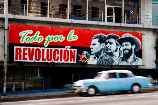 Havana, Cuba - May 13, 2010: Classic 50's American made car passes under iconic billboard promoting the ongoing Communist revolution in the La Rampa neighborhood. The billboard depicts Julio Mella, Che Guevara, and Camilo Cienfuegos, three key figures in the country's Communist revolution.