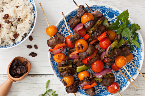 An overhead close up shot of several lamb kabobs on a plate and a bowl of rice with raisins.