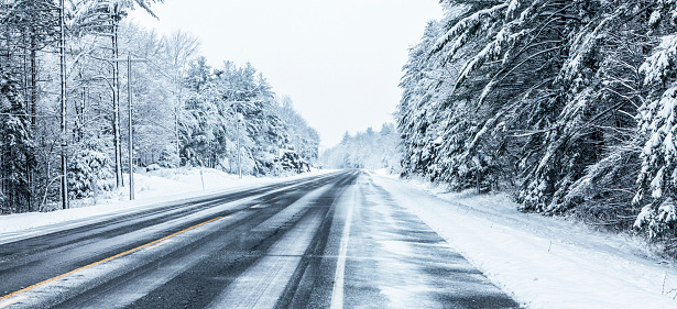 A long, remote, straight stretch of of rural highway NY State Route 3 in the Adirondacks Mountains region near the northern New York State hamlet of Cranberry Lake during a strong winter blizzard snow storm. The road is lined with forest branches bent under the weight of the heavy, wet snow.