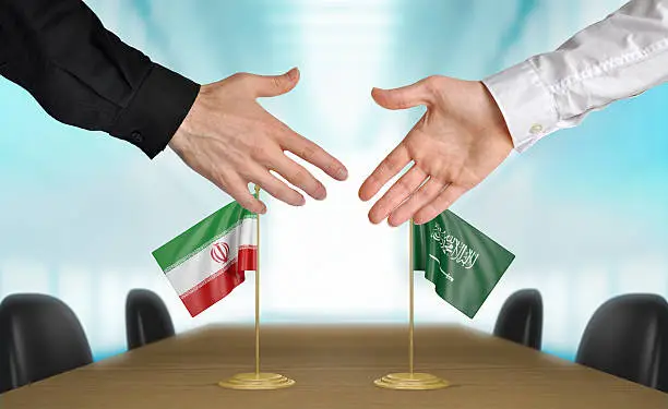 Two diplomats from Iran and Saudi Arabia extending their hands for a handshake on an agreement between the countries.