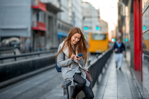 Young woman in casual gray clothes using smartphone sitting near the street on the gray city background