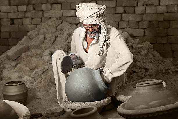 Traditional Indian Potter Sitting wearing white Kurta-Pajama & Turban which is Traditional Dress for Men in North India. He is hitting the Clay Pots to giving it Proper Round Shape on Raw Brick wall in the Background.