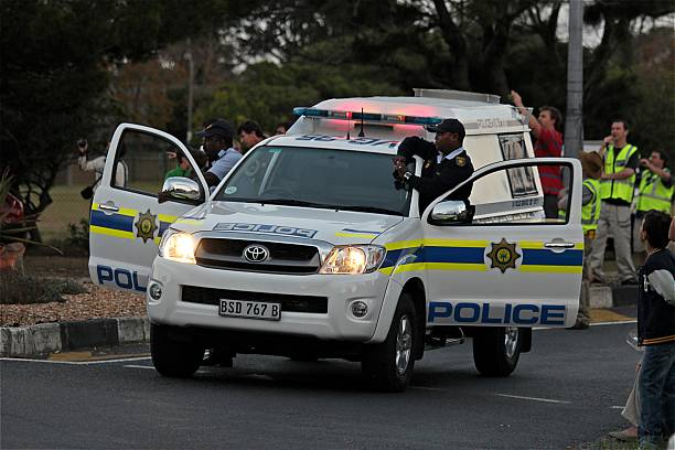 Cape Town, South Africa - April 13th, 2011: A standard patrol vehicle for the South African Police is stopped blocking a road with its doors open and lights on at dusk. Two uniformed police officers stand behind each door watching a public demonstration to the left. Behind on the rights stands some members of a local neighborhood watch in yellow reflective vests.