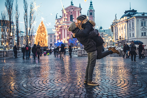 Portrait of young couple in the old city center on cold winter day. Man is holding woman in the air while they are kissing. They are happy together. People and city with christas lights in back, defocused.