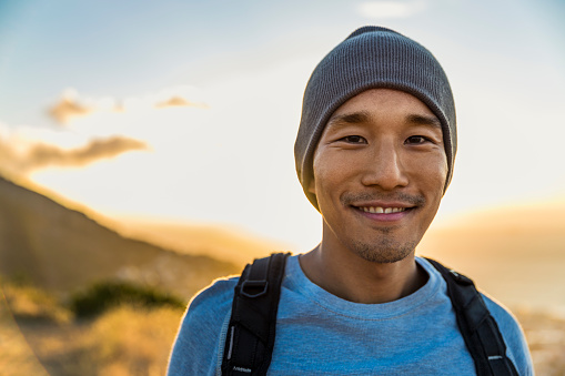 A photo of smiling male backpacker on hill. Happy traveler is wearing knit hat. Portrait of man is enjoying vacation against sky during sunset.