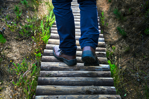 A photo of hiker walking on boardwalk. Low section of man is in forest. Young male is wearing shoes while moving on wood. He is hiking during weekends.