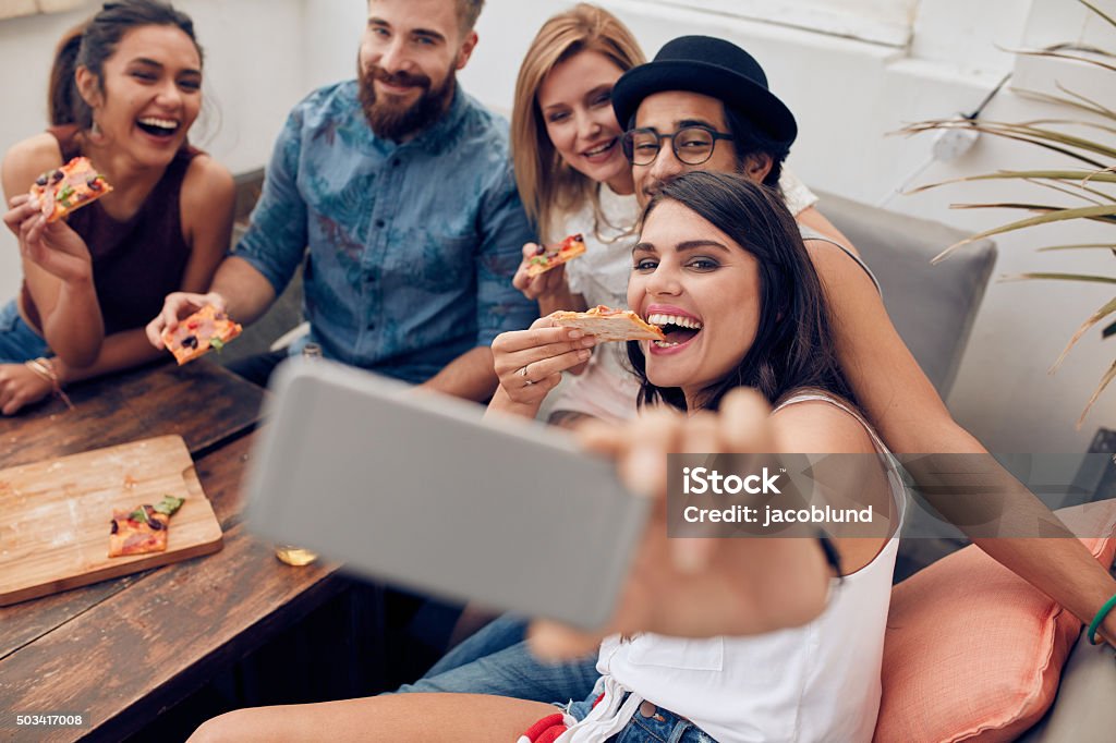 Young people taking a selfie while eating pizza Group of multiracial young people taking a selfie while eating pizza. Young woman eating pizza her friends sitting around during a party. Pizza Stock Photo